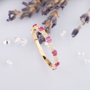 May include: A delicate gold band ring with alternating pink and white gemstones. The ring is set with small square-shaped stones.