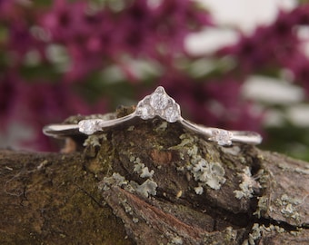 May include: A delicate silver ring with a cluster of small diamonds set in a curved, v-shaped design. The ring is resting on a piece of bark with a mossy texture.