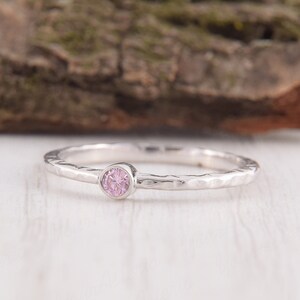 May include: A silver ring with a hammered band and a small, round, pale pink gemstone. The ring is set on a white surface, with a blurred wooden background. The ring's design is simple and elegant, suitable for everyday wear.