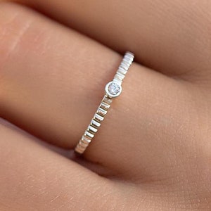 Op de afbeelding: Een zilveren ring met een kleine, heldere edelsteen in het midden. De band van de ring heeft een getextureerd, geribbeld ontwerp.