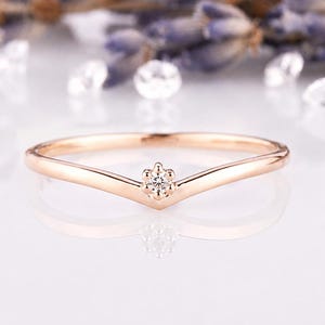 May include: A rose gold V-shaped ring featuring a single small round brilliant-cut diamond set in a six-prong setting at the center point of the band. The metal has a polished finish and the band curves into a chevron shape.
