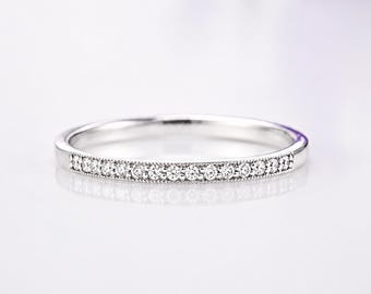 May include: A thin silver-coloured band ring featuring a row of small, clear, round-cut stones set along the front half. The metal band has a smooth, polished finish with delicate milgrain detailing bordering the stones on both sides.