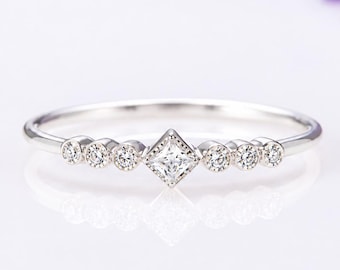 May include: A silver-toned ring featuring a central princess-cut stone set in a diamond-shaped bezel, flanked by three small round stones on each side. The band is thin and polished, showcasing a delicate arrangement of clear, sparkling stones.