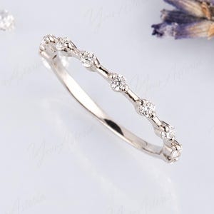 Könnte beinhalten: Ein dünner Ring aus silberfarbenem Metall mit einer Reihe kleiner, runder, klar facettierter Steine, die entlang der oberen Hälfte des Bandes gefasst sind. Die Steine sind gleichmäßig in einzelnen Fassungen angeordnet und bilden eine feine Linie.