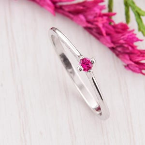 Könnte beinhalten: Ein dünner, polierter Ring aus silberfarbenem Metall mit einem einzelnen, kleinen, rund geschliffenen, leuchtend pinkfarbenen Edelstein in einer Vier-Krappen-Fassung. Die Ringschiene ist glatt und schmal und hat eine glänzende Oberfläche.