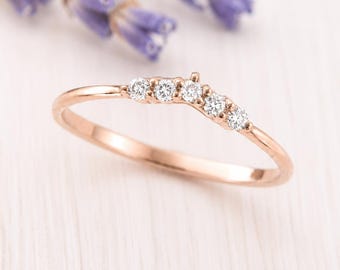 May include: A rose gold ring featuring a delicate V-shaped band set with five small, round, clear stones arranged in a row along the top curve. The metal has a smooth, polished finish, and the design follows a subtle chevron-inspired silhouette.