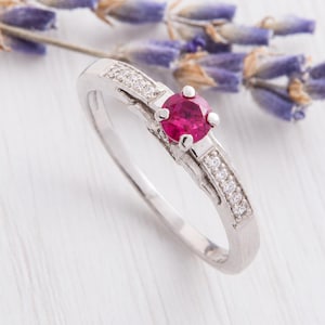 Könnte beinhalten: Ein silberfarbener Ring mit einem runden, leuchtend pinkfarbenen Mittelstein in einer Vier-Krappen-Fassung. Die Ringschiene ist auf beiden Seiten mit kleinen, klaren, rund geschliffenen Steinen besetzt, was für ein poliertes und glänzendes Aussehen sorgt.
