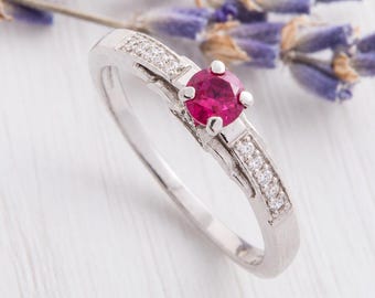 May include: A silver-toned ring featuring a round, vibrant pink centre stone held by a four-prong setting. The band is accented with small, clear, round-cut stones embedded along both sides of the shank, creating a polished and reflective appearance.