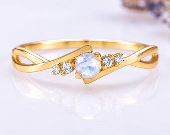 May include: A gold ring featuring a central round iridescent moonstone flanked by two small clear round stones on each side. The band has a crossover design that tapers toward the center setting, creating a delicate and symmetrical appearance.