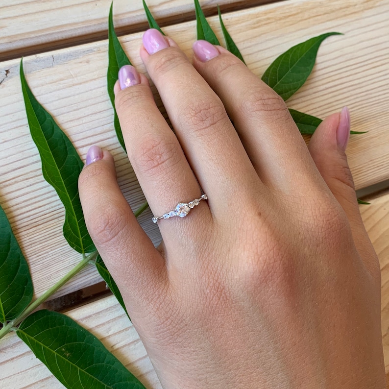 May include: A delicate silver ring with a cluster of small, clear gemstones. The ring is set on a finger with a light pink nail polish.