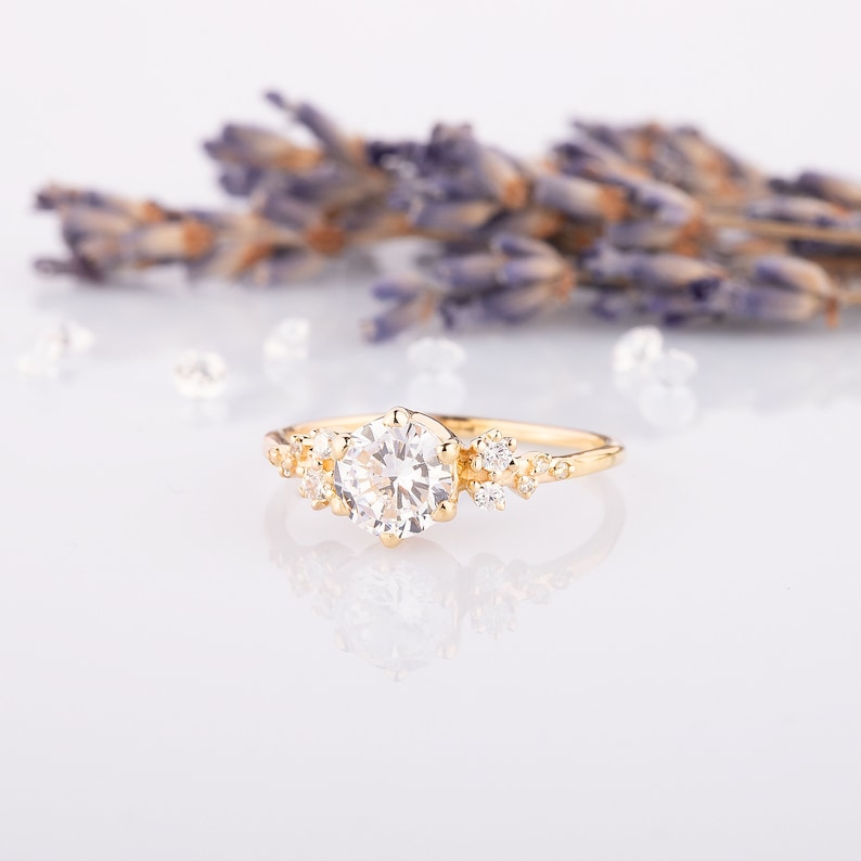 K&ouml;nnte beinhalten: Ein zarter goldener Verlobungsring mit einem gro&szlig;en runden Mittelst&uuml;ck und kleineren Akzent-Steinen. Der Ring liegt auf einem wei&szlig;en Hintergrund mit Lavendelbl&uuml;ten.