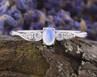 Op de afbeelding: Een zilverkleurige ring met een centrale ovale iriserende maansteen cabochon, geflankeerd door twee kleine heldere rondgeslepen stenen aan elke kant, gezet in een gepolijste metalen band die licht buigt rondom de centrale zetting.