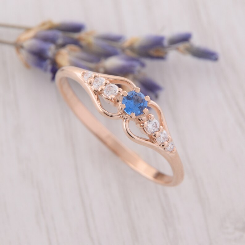 May include: A delicate rose gold ring with a blue gemstone surrounded by smaller white gemstones. The ring features a delicate heart-shaped design.