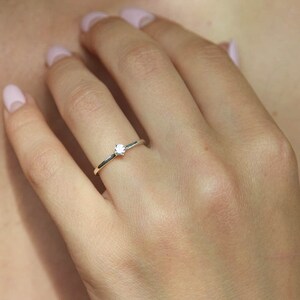 Pu&ograve; includere: Un delicato anello d'argento con una piccola gemma trasparente al centro. L'anello ha una fascia sottile e un design semplice ed elegante. L'anello &egrave; mostrato su un dito.