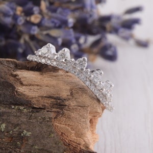 May include: A silver-toned crown-shaped ring featuring a band encrusted with numerous small, clear, sparkling stones and larger round stones set along the top edge, creating a jagged, elevated silhouette that resembles a royal tiara design.