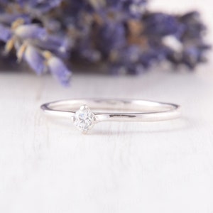 Pu&ograve; includere: Un delicato anello d'argento con un piccolo diamante a taglio quadrato al centro. La fascia dell'anello &egrave; sottile e lucida, riflettendo la luce. Lo sfondo presenta fiori viola sfocati, creando un'estetica romantica ed elegante.