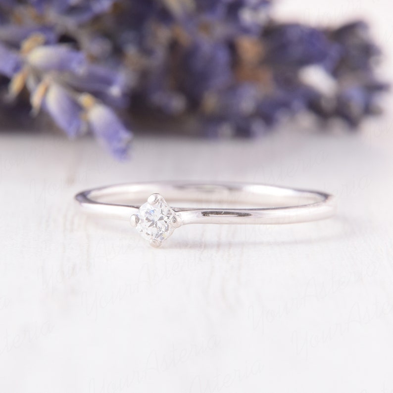Pu&ograve; includere: Un delicato anello d'argento con un piccolo diamante a taglio quadrato al centro. La fascia dell'anello &egrave; sottile e lucida, riflettendo la luce. Lo sfondo presenta fiori viola sfocati, creando un'estetica romantica ed elegante.