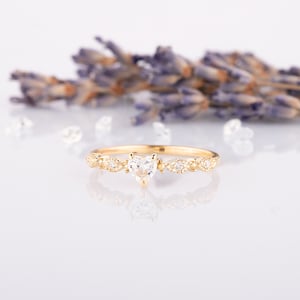 May include: A gold ring with a heart-shaped clear gemstone at the center. The band features a delicate design with small, round gemstones and leaf-like accents. The ring is set against a soft, white background with lavender sprigs.