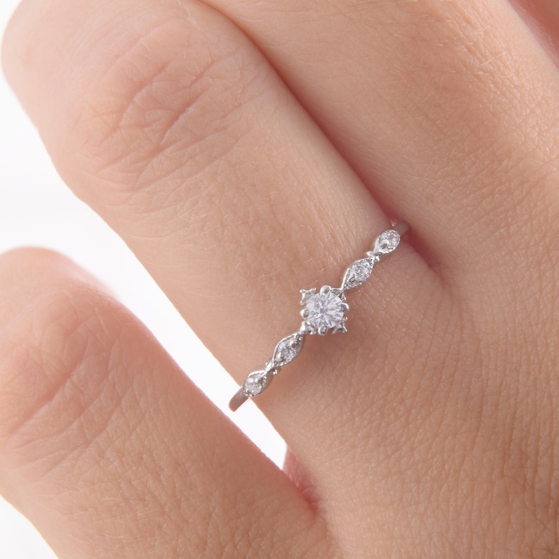 May include: A delicate silver ring with a small round diamond in the center and smaller diamonds set in a curved design along the band.