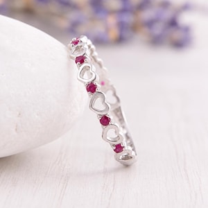 May include: A silver-toned ring featuring an alternating pattern of open heart shapes and small, round, vibrant pink stones. The band is constructed with a repeating sequence of these heart motifs and circular settings, creating a continuous design.