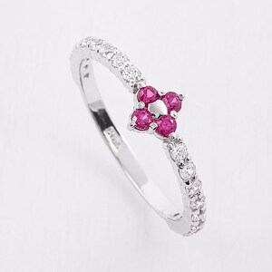 May include: A silver-toned ring featuring a central flower-shaped cluster of four round-cut vibrant pink stones with a small metallic dot in the center, flanked by a row of clear, round-cut white stones set along the band on both sides.