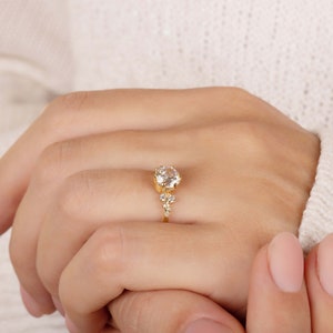 K&ouml;nnte beinhalten: Ein goldener Ring mit einem gro&szlig;en, klaren, runden Edelstein und kleineren Edelsteinen auf der Ringschiene.