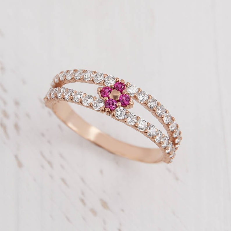Puede incluir: Un delicado anillo de oro rosa con una doble banda de piedras blancas brillantes. Un peque&ntilde;o grupo de piedras rosas en forma de flor se encuentra en el centro del anillo.