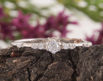 May include: A delicate silver ring with a single, round, clear gemstone surrounded by smaller, clear gemstones. The ring is set on a rustic, brown background.