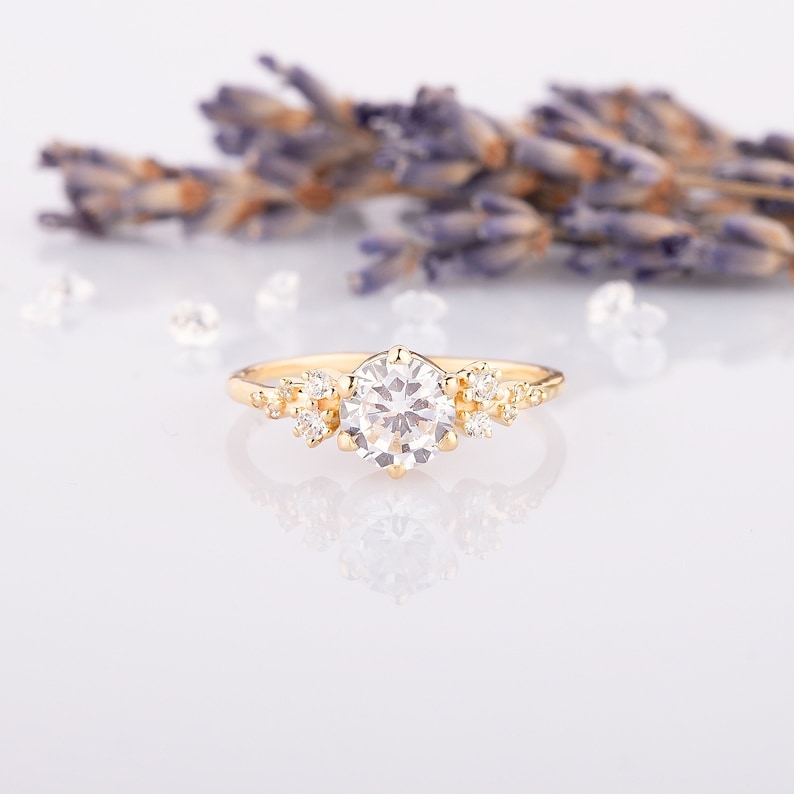 K&ouml;nnte beinhalten: Ein zarter goldener Verlobungsring mit einem gro&szlig;en runden Mittelst&uuml;ck und kleineren Akzent-Steinen. Der Ring liegt auf einem wei&szlig;en Hintergrund mit Lavendelbl&uuml;ten.