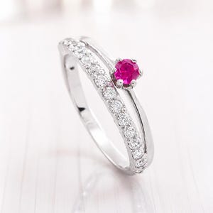 May include: A silver-toned ring featuring a split-shank design. One side of the band is adorned with a row of small, clear, round-cut stones, while the other side is a smooth, plain band topped with a single, vibrant, round-cut fuchsia-pink stone.