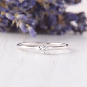Pu&ograve; includere: Un delicato anello d'argento con una singola gemma rotonda e trasparente. L'anello &egrave; posto su una superficie bianca, con uno sfondo sfocato di fiori viola. La fascia dell'anello &egrave; sottile e lucida, e la gemma &egrave; tenuta da quattro griffe.