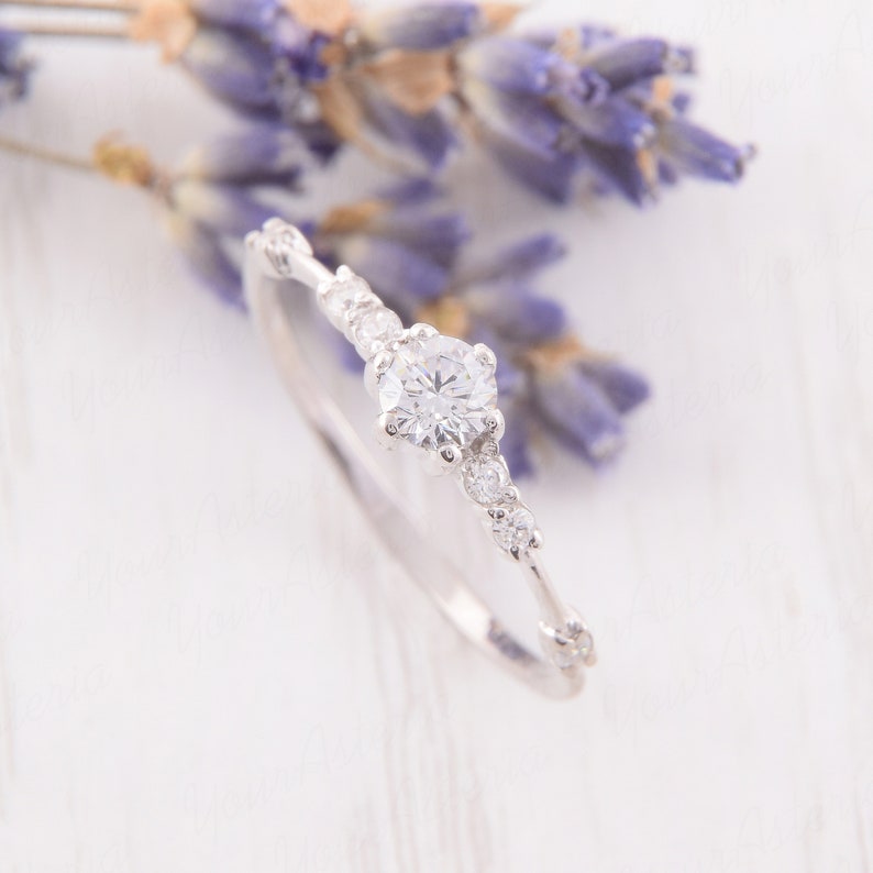May include: A delicate silver ring with a single round diamond and smaller diamonds set along the band. The ring is on a white background with lavender flowers.
