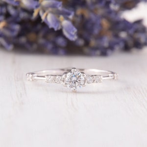 May include: A delicate silver engagement ring with a single round diamond surrounded by smaller diamonds. The ring is set on a white wooden surface.