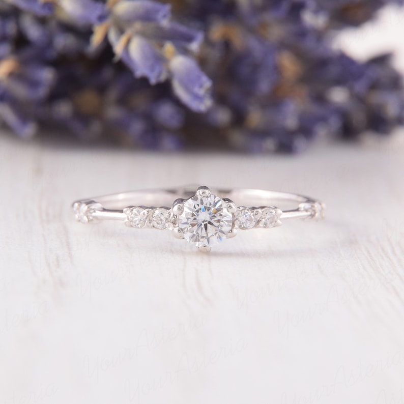 May include: A delicate silver engagement ring with a single round diamond surrounded by smaller diamonds. The ring is set on a white wooden surface.