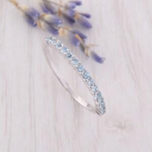 Op de afbeelding: Een delicate zilveren bandring met een rij lichtblauwe edelstenen. De ring ligt op een witte houten achtergrond.