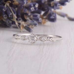 May include: A delicate silver ring with a small round diamond in the center and smaller diamonds on each side. The ring is set on a white wooden surface.