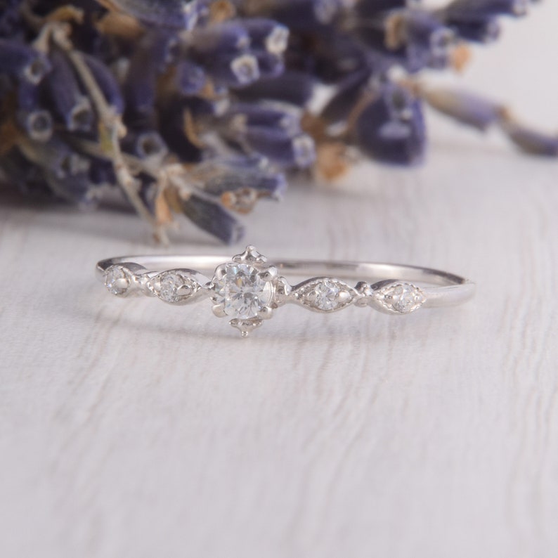 May include: A delicate silver ring with a small round diamond in the center and smaller diamonds on each side. The ring is set on a white wooden surface.