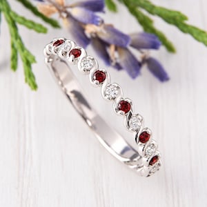 Op de afbeelding: Een zilverkleurige ring met een afwisselend patroon van ronde, dieprode edelstenen en heldere, sprankelende witte stenen, gezet in een doorlopend, vloeiend metalen ontwerp dat zorgt voor een delicate, onderling verbonden uitstraling.