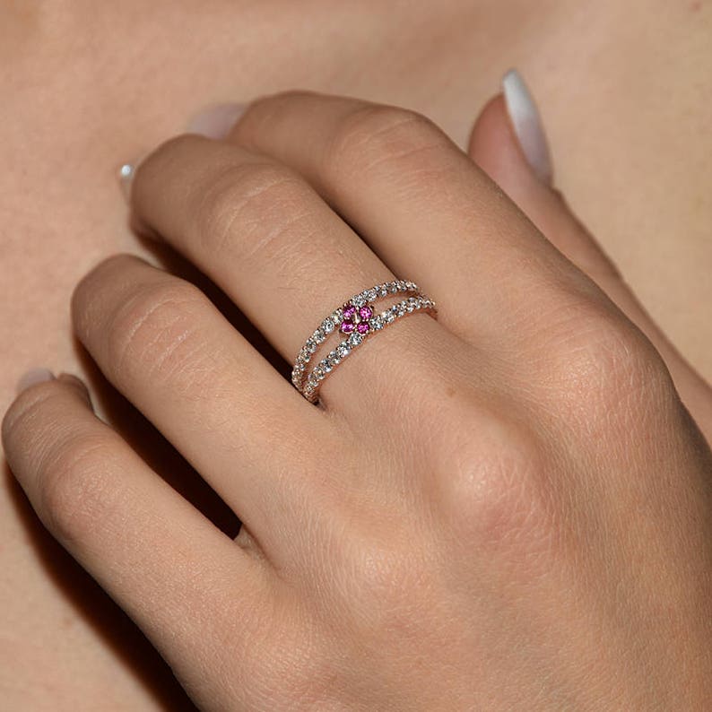 Puede incluir: Un delicado anillo con una doble banda de piedras transparentes y una flor central con piedras rosas.