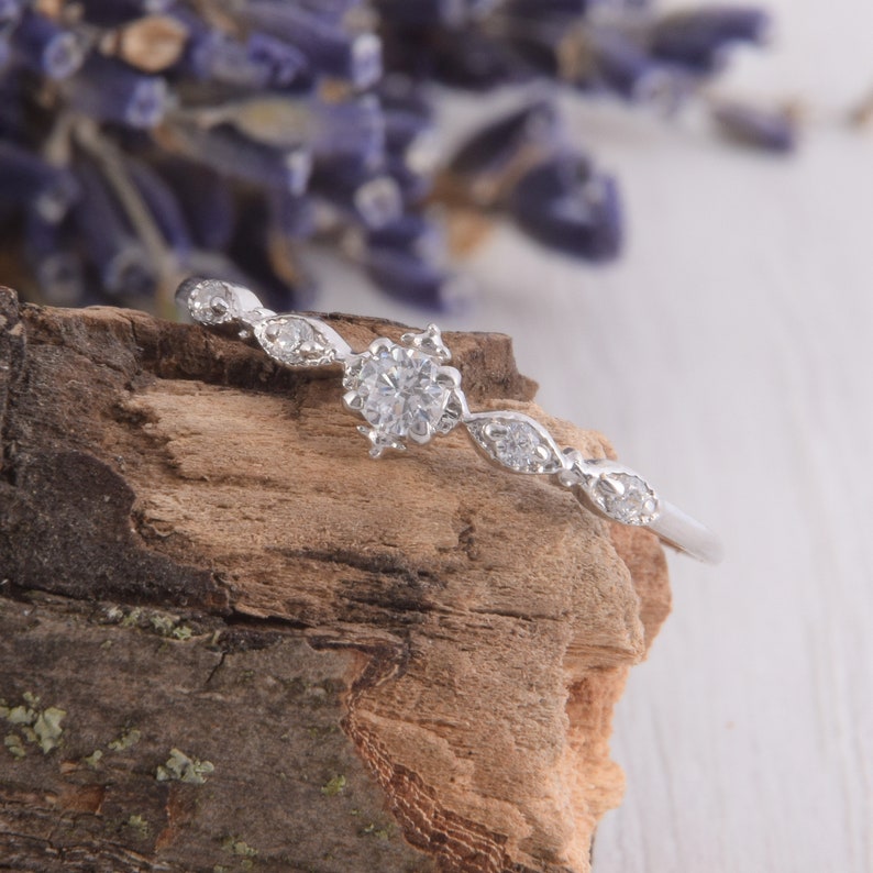 May include: A delicate silver ring with a single round-cut diamond surrounded by smaller diamonds. The ring is set on a piece of weathered wood.