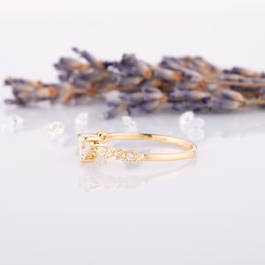 May include: A delicate gold ring with a central, clear gemstone and smaller accent stones. The band features a decorative pattern with tiny, sparkling embellishments. The ring is set against a soft, white background with lavender sprigs.