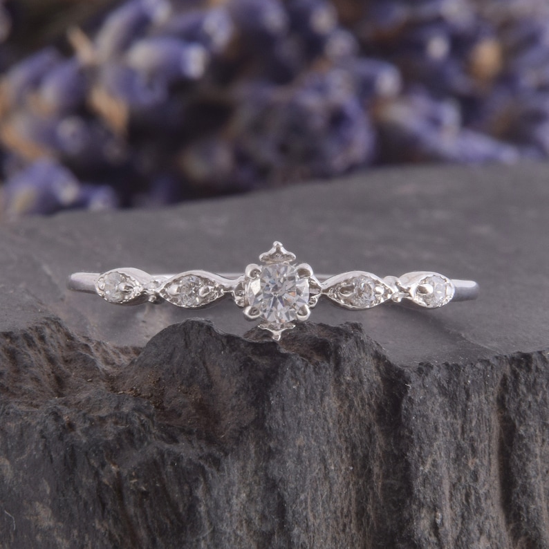 May include: A delicate silver ring with a single round-cut diamond in the center and smaller diamonds on each side. The ring has a vintage-inspired design.