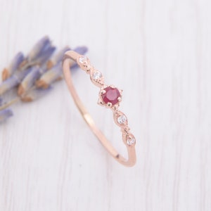May include: A delicate rose gold ring with a single red ruby gemstone surrounded by smaller white diamonds. The ring has a simple, elegant design.
