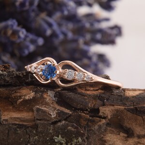 May include: A delicate rose gold ring with a blue sapphire center stone and smaller clear stones set in a heart-shaped design.