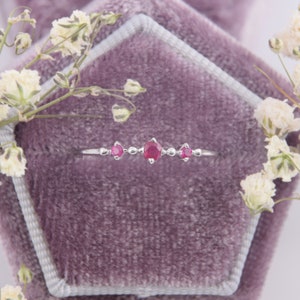 May include: A delicate silver ring with three small ruby stones set in a row. The ring is displayed on a purple velvet cushion.