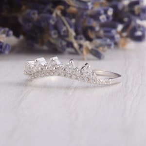 May include: A silver-toned crown-shaped ring featuring a curved band adorned with multiple small, clear, sparkling stones. The design includes a series of pointed, elevated settings along the top edge, creating a distinct tiara-like silhouette.