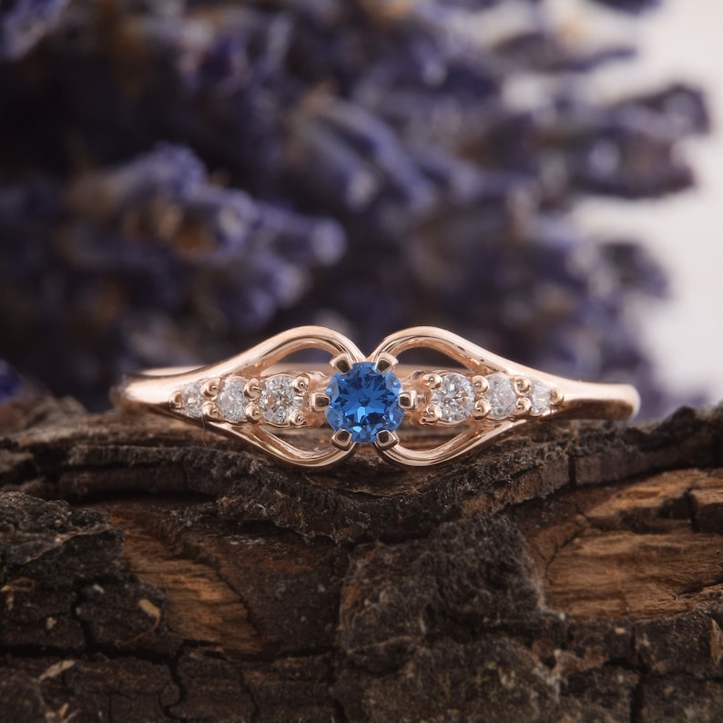 May include: A rose gold ring with a blue gemstone surrounded by smaller white gemstones. The ring has a delicate, openwork design.