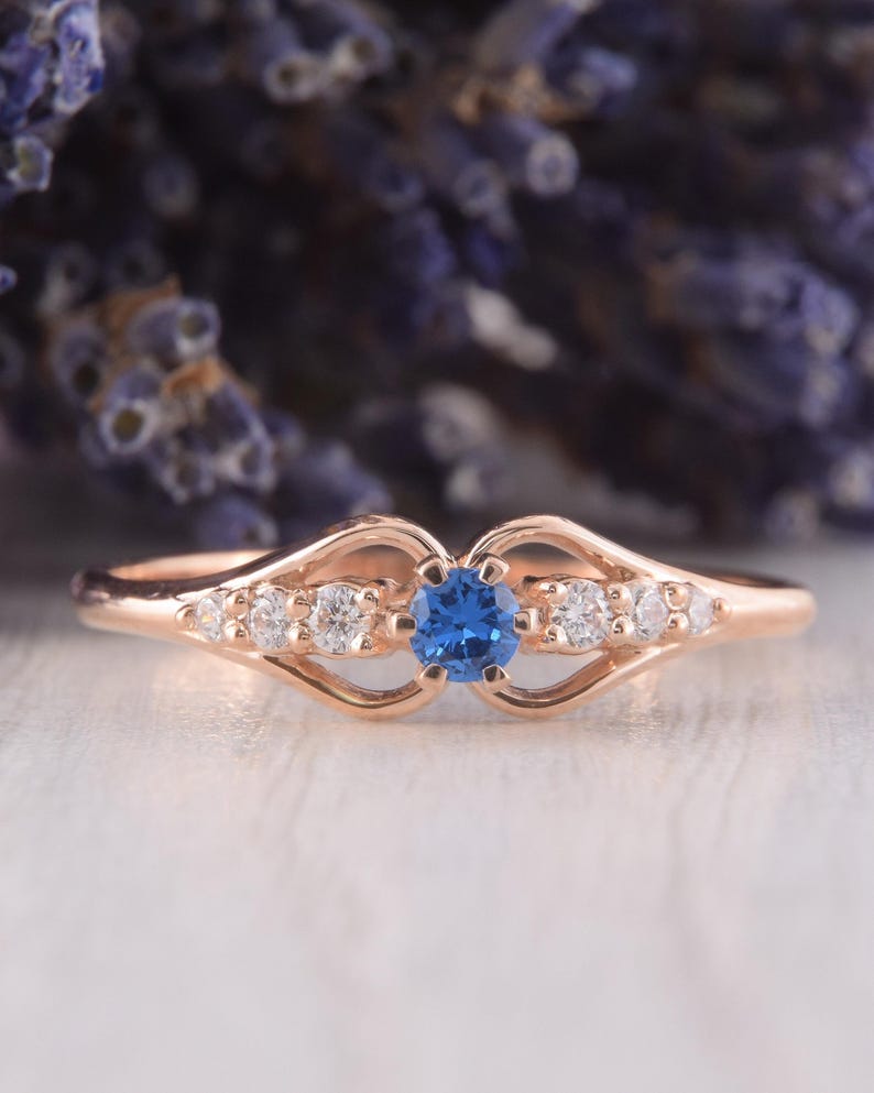 May include: A rose gold ring featuring a central round blue gemstone flanked by three small clear stones on each side. The band has a split design that curves around the stones, creating an open, symmetrical shape along the finger.