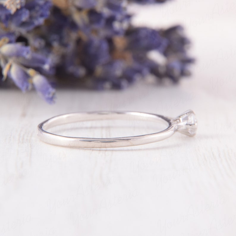Pu&ograve; includere: Un delicato anello d'argento con una singola gemma trasparente. L'anello &egrave; posto su una superficie bianca, con uno sfondo sfocato di fiori viola. Il gambo dell'anello &egrave; sottile e lucido, e la gemma &egrave; rotonda e rialzata.
