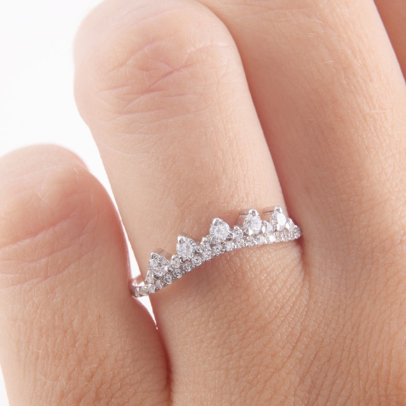 May include: A silver-toned ring featuring a crown-shaped design with a row of small, clear, round-cut stones along the band and five larger, prominent, clear, round-cut stones set in a pointed, elevated arrangement across the top of the ring.